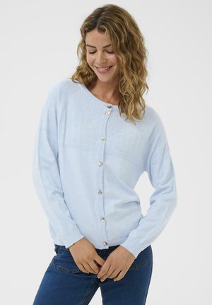 Kvinde med bølget lysbrunt hår iført en lyseblå knapcardigan og mørkeblå jeans, smilende med lukkede øjne mod en ensfarvet baggrund.