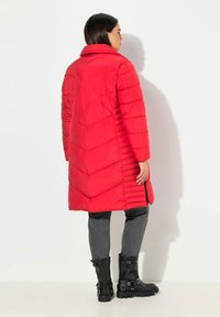 Piumino rosso con cuciture a zigzag, colletto alto e maniche lunghe. Abbinato a stivaletti neri alla caviglia e pantaloni grigi.