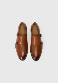 Zapatos de cuero marrón tipo slip-on con dos hebillas, con un acabado liso, puntera redondeada y detalles de costura horizontal. Suela plana negra.