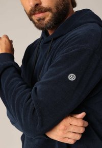 Haut en velours côtelé bleu marine avec une finition texturée, capuche à cordon réglable et écusson rond sur la manche gauche.