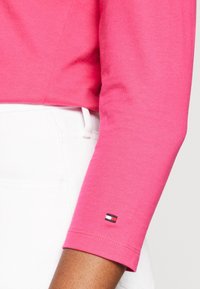 Camicia a maniche lunghe rosa con texture a costine, dotata di un patch del logo ricamato sul polsino. Abbinata a pantaloni bianchi.
