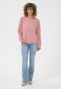 Light pink long-sleeve blouse with textured diamond patterns, round neckline, and gathered sleeves, paired with light blue flared jeans and beige shoes.