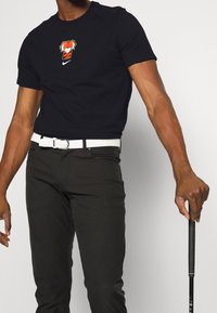 Svart bomullströja med en röd och orange grafik på bröstet, kombinerad med mörka byxor och ett vitt flätat bälte. Golfklubba i handen.