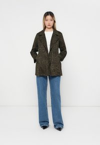 Olivgrön dubbelknäppt kappa med leopardmönster, sidofickor, slagkragar och slät textur, kombinerad med blå vida jeans.