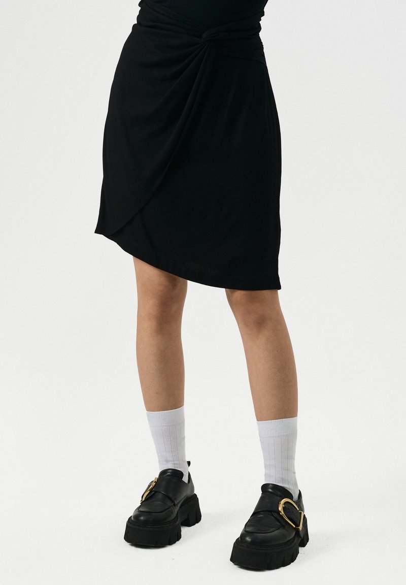 Persoon in een zwarte asymmetrische rok, witte geribde crew-sokken en zwarte stevige loafers met grote gouden gespen op een witte achtergrond.