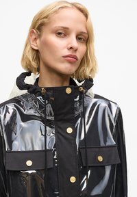 Chubasquero negro transparente con capucha, botones dorados, detalles en tejido negro y bolsillos delanteros. Textura suave y diseño elegante.