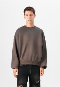 Sweatshirt castanho de grandes dimensões feito de tecido macio, com um decote redondo, punhos canelados e uma bainha com acabamento franzido. Combinado com jeans pretos desgastados.