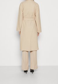 Abrigo de lana beige con cintura ceñida, mangas largas y silueta recta, combinado con pantalones de pierna ancha beige y tacones negros.