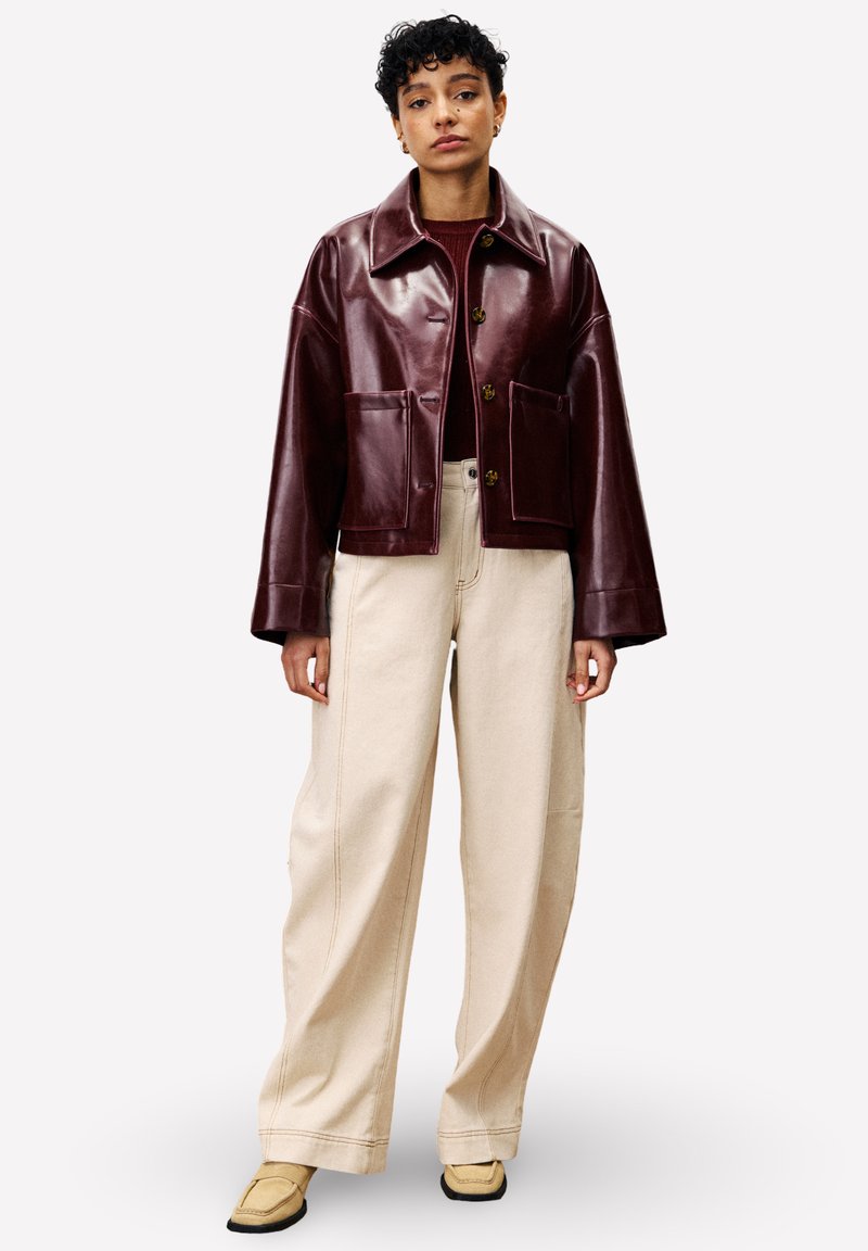 Bordeaux leren jack met een kraaghalslijn, twee voorzakken en gouden knopen, gecombineerd met brede beige broeken en tan schoenen.