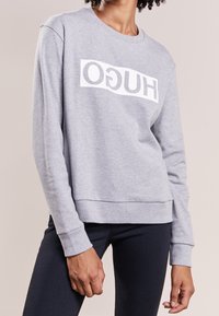 Sudadera gris hecha de material suave, con un destacado logo blanco de "HUGO". El diseño incluye un cuello redondo y puños de canalé.