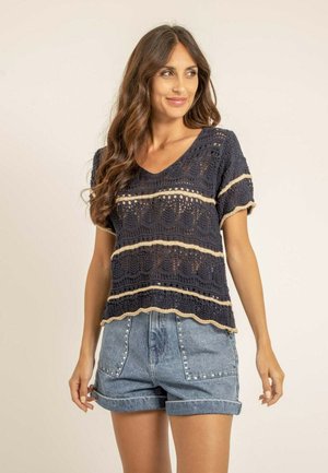 Femme aux longs cheveux ondulés portant un haut court en crochet bleu marine avec des rayures beiges et un short en jean bleu clair avec des détails cloutés.