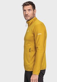 Gelbe Zip-Jacke aus strukturiertem Material, mit Stehkragen, seitlicher Tasche und einem Logo auf der Schulter.