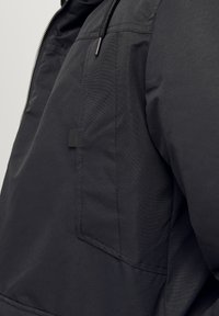 Veste en tissu noir avec une fermeture éclair et une poche extérieure. Présente une finition texturée et un cordon de serrage attaché à la capuche.
