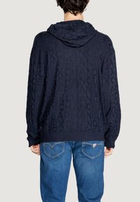 Felpa con cappuccio blu navy caratterizzata da un motivo testurizzato del nome del marchio. Ha una vestibilità rilassata con polsini e orlo a coste. Abbinata a jeans blu.