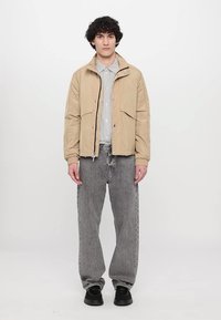Beige bomberjakke med glidelås foran, to brystlommer og strikkede mansjetter, parret med grå baggy jeans og svarte sneakers.