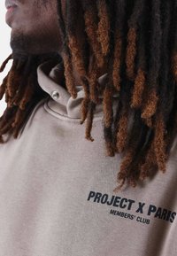Sudadera beige con cuello alto, con texto negro "PROJECT X PARIS" y "MEMBERS' CLUB". Tejido suave, con detalles de costura visibles.