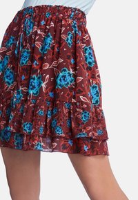 Jupe fleurie avec un fond bordeaux, présentant des fleurs bleues et rouges, une ceinture élastique et des volants superposés pour une texture ajoutée.