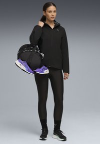 Veste zippée noire, leggings noirs, chaussures noires avec des accents teal, et un sac de sport noir contenant des baskets violettes, sur un fond gris.