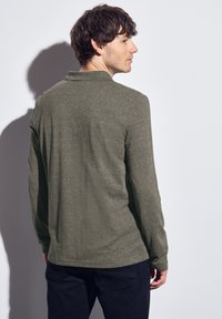 Polo à manches longues vert olive en tissu texturé, avec un détail de col et un ourlet droit, porté avec un pantalon sombre, montré de dos.