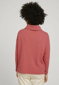 Przeoversizowy, teksturowany sweter typu pullover w kolorze przydymionego różu z szerokim kołnierzem i długimi rękawami, zestawiony z jasnymi spodniami.