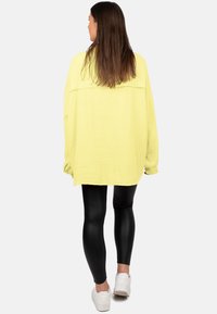 Camicia gialla oversize con texture e maniche lunghe, abbinata a leggings neri e sneaker bianche, vista da dietro.