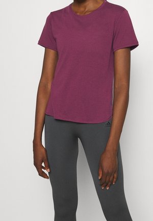 Osoba nosící krátké fialové tričko a tmavě šedé legíny Adidas stojí před jednoduchým světlým pozadím.