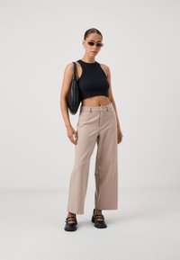 Svart kort topp, beige vida byxor, svarta platta skor med remmar och en svart handväska. Modellen står mot en vit bakgrund.