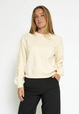 Junge Frau mit langen blonden Haaren, die einen cremefarbenen Pulli mit Spitzenakzenten an den Ärmeln und schwarzen Hosen trägt, steht vor einem schlichten weißen Hintergrund.