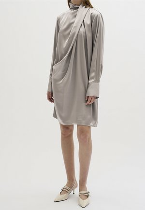 Kobieta ubrana w luźną, długorękawową, jasnoszarą sukienkę z satyny z drapowanym detalem, zestawioną z kremowymi szpilkami z podwójnymi paskami i ostrym noskiem.