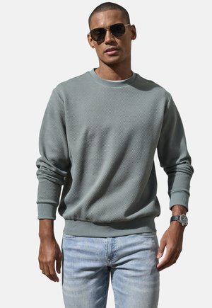 Mann mit dunkler Sonnenbrille, grauem strukturiertem Langarmpullover, hellblauer Jeans und Armbanduhr vor unifarbenem Hintergrund.