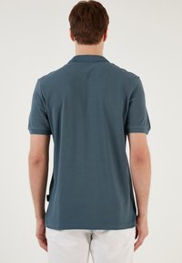 Paita polo dalam warna biru lembut, lengan pendek, tekstur halus, dengan kerah bulat dan detail jahitan yang halus di belakang.