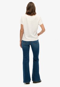 Hvid kortærmet t-shirt med rund halsudskæring, kombineret med blå flared jeans med baglommer og sømdetalje. Sort fodtøj synligt.