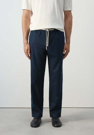 Uomo che indossa pantaloni blu navy con coulisse bianca e scarpe nere, in piedi davanti a uno sfondo chiaro uniforme.