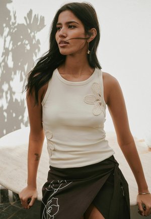 Woman with long dark hair wears sleeveless beige top with crochet flowers and black skirt with white floral embroidery, standing by round table.