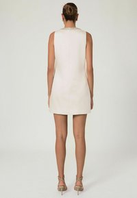 Bezrukávové, po kolená siahajúce šaty z jemného krémového materiálu s hladkým povrchom. Obsahujú ozdobu na výstrihu, jednoduchý strih a zips na zadnej strane.