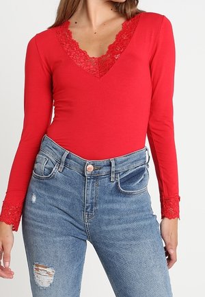 Femme portant un haut rouge à manches longues avec bordure en dentelle au niveau du col et des poignets, associé à un jean bleu déchiré à taille haute.