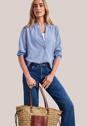 Femme portant un chemisier rayé bleu et un jean, tenant un sac cabas en paille tissée avec des poignées en cuir marron et une poche avant.