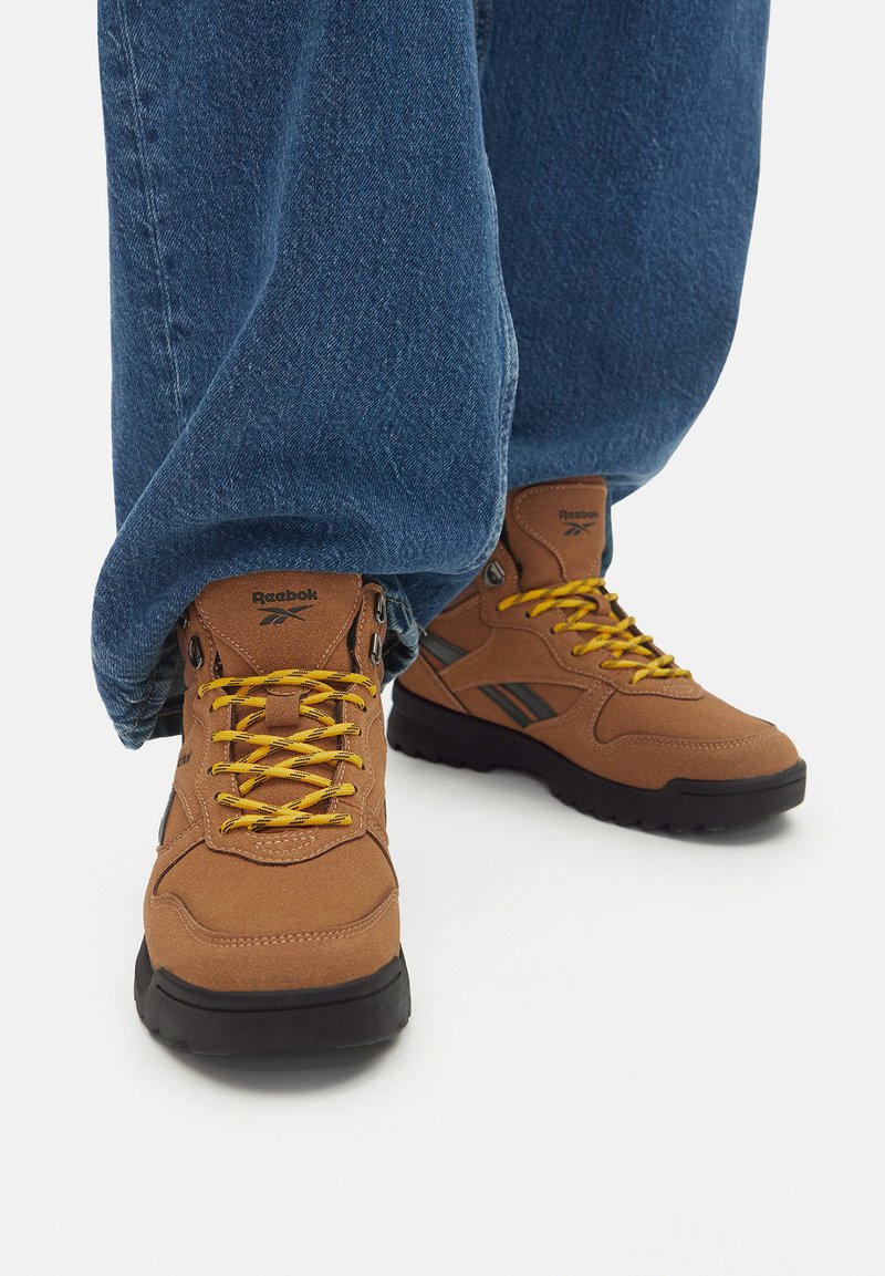 Hnedé turistické topánky so žltými šnúrkami, z semišového materiálu, s čiernymi gumovými podrážkami a logom Reebok; skombinované s voľnými modrými denimovými nohavicami.