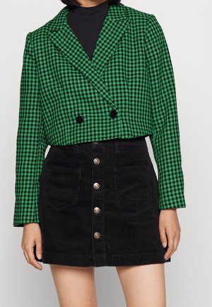 Femme portant un blazer court à motif pied-de-poule vert et noir sur un col roulé noir, associé à une jupe en velours côtelé noire avec boutons sur le devant.