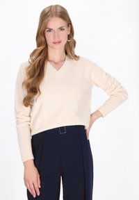 Pulover crem cu decolteu în V, dintr-un tricot moale, cu manșete ondulate, asortat cu pantaloni bleumarin cu talie înaltă și cercei rotunzi din argint. Textură netedă, design ajustat.
