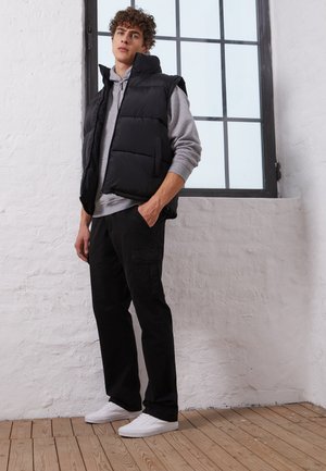 Schwarze gepolsterte ärmellose Weste über einem grauen Hoodie, kombiniert mit schwarzen Cargo-Hosen und weißen Sneakers. Glattes Material, kastiger Schnitt, minimalistisches Design.