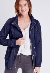 Giacca leggera blu navy con chiusura a zip, cordino regolabile e dettagli con bottoni. Indossata sopra un top bianco a maglia testurizzata.