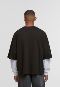 Mørkebrunt oversized t-shirt, korte ærmer, rund hals, blødt bomuldsmateriale, viser bagsiden med grå jeans og hvid undertrøje synlig.