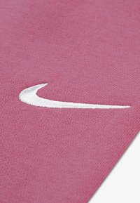 Nike'i logo tekstureeritud roosa kanga taustal; logo on valge ja tikkitud, kujutab kõverat, lennukujulist disaini.