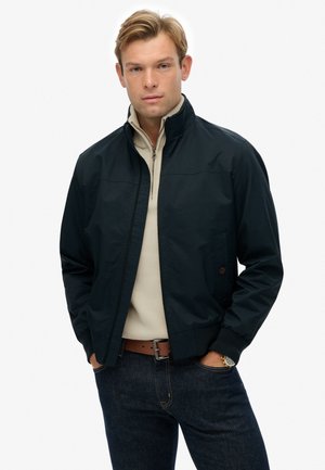 Navy bomber jacket with a high collar, front zipper, and elastic cuffs. Worn over a beige light sweater, paired with dark jeans and a brown belt.