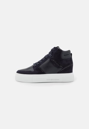 Zapatilla negra de caña alta con paneles de ante y cuero, detalles con tachuelas negras, cordones en blanco y negro, y suela gruesa de goma blanca.