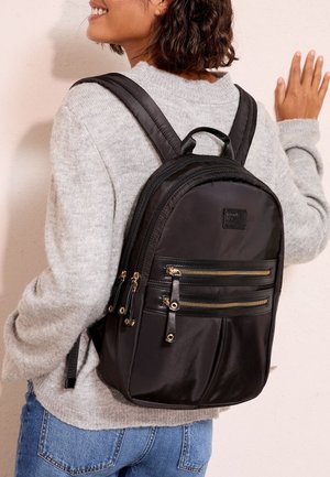 Donna con maglione grigio e jeans blu con uno zaino nero con più scomparti con zip e spallacci imbottiti.