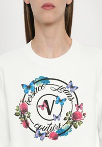 Felpa bianca con grafica floreale e farfalle colorate, con un logo circolare nero che recita "Versace Jeans Couture" centrato sul petto.