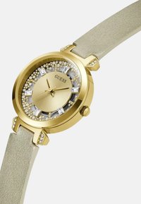 Orologio dorato con accenti di cristalli scintillanti, cinturino in pelle beige, quadrante rotondo e lunetta circolare con un centro testurizzato e indicatori delle ore.