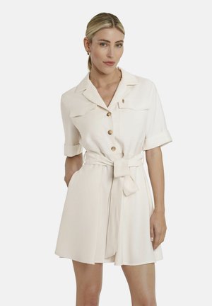 Frau mit blonden Haaren, die ein cremefarbenes, kurzärmeliges Hemdkleid mit Taillengürtel und hochgekrempelten Manschetten trägt, steht vor einem schlichten Hintergrund.